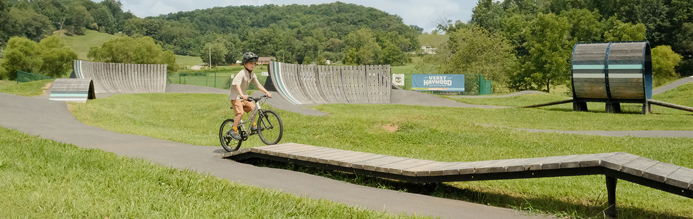 Raccoon Creek Bike Park - Young Boy biking toward a wooden ramp at a park with mountains in the background