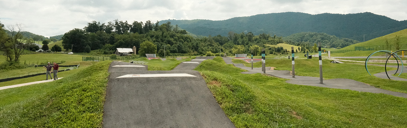Raccoon Creek Bike Park - Showing pump track with hilly berms for bicycle jumps. two men walk on the trail surrounding the pump track