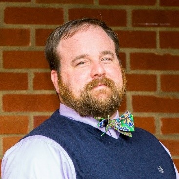 Tyrus Clayton - Man with brown hair and beard, wearing collared shirt and vest with an expressive bowtie